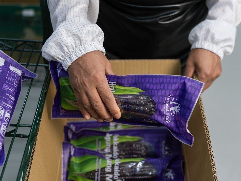 Hands holding a finished Natural Fare purple sweet corn pack over a Natural Fare carton of packs